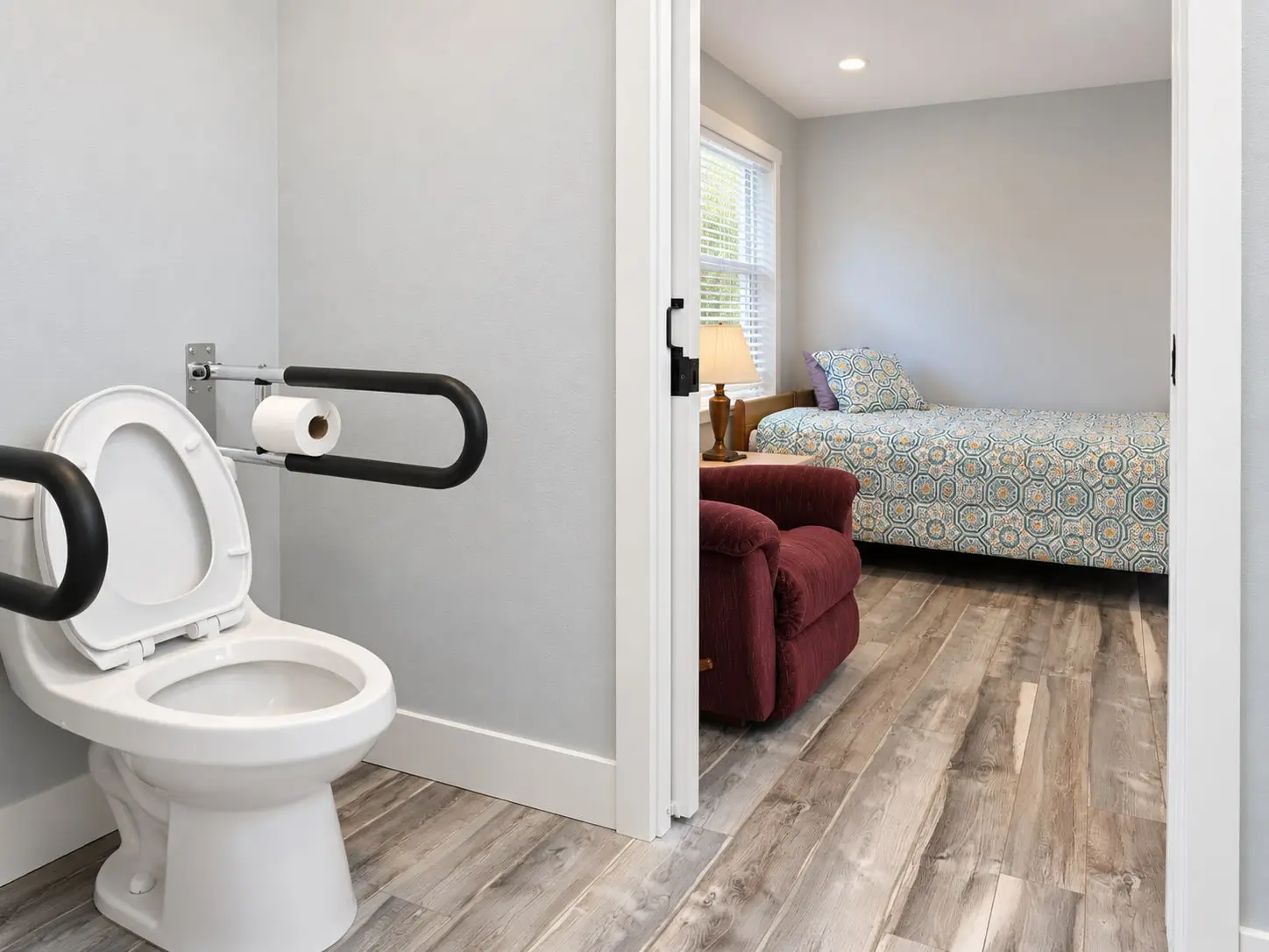 Toilet with grab bars beside a doorway into a bedroom
